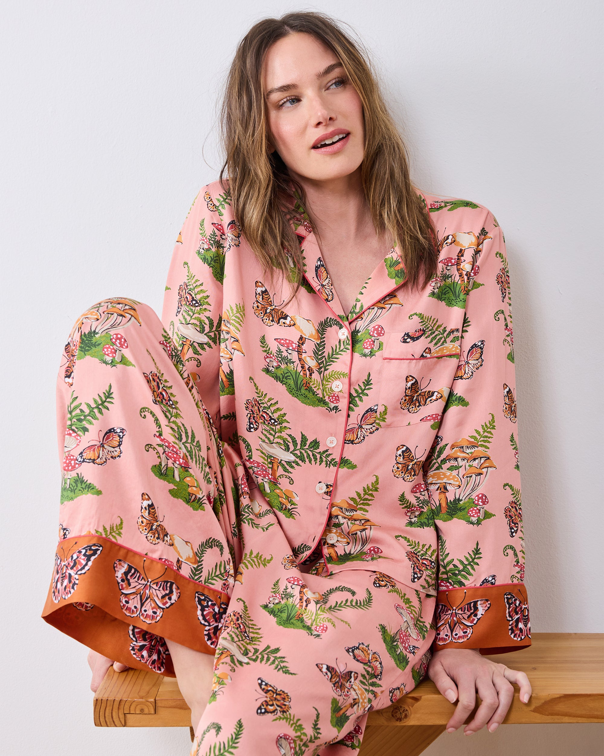 Model seated on a wooden bench wearing the pink Butterfly Forager pajama set with a button-front top, chest pocket, and wide-leg pants.