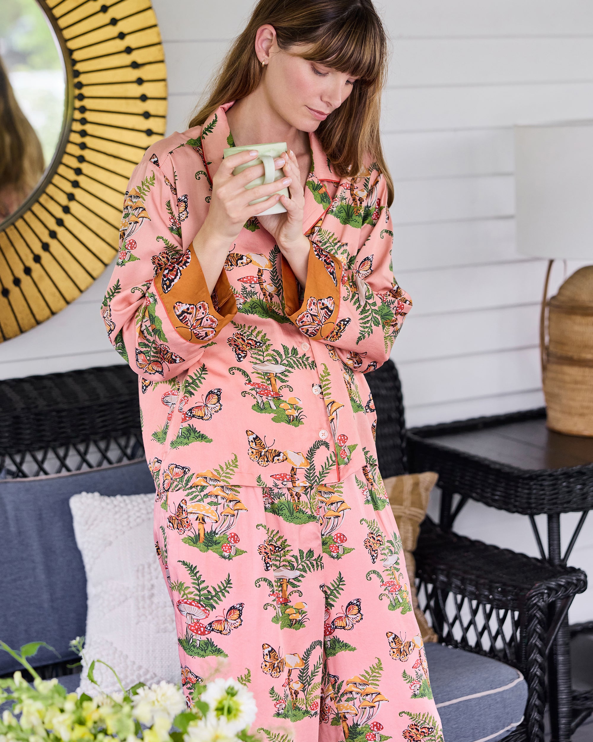 Person wearing the Butterfly Forager rosewood Soho pajama set, with a button-front top and wide-leg pants printed with butterflies, mushrooms, and ferns, holding a cup.