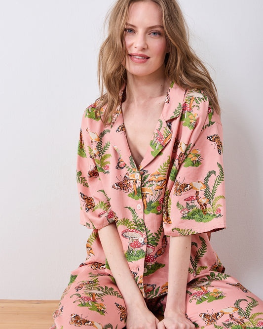 Seated view of a rosewood pajama set with a butterfly, mushroom, and fern print, showing the notched collar and button-front top.