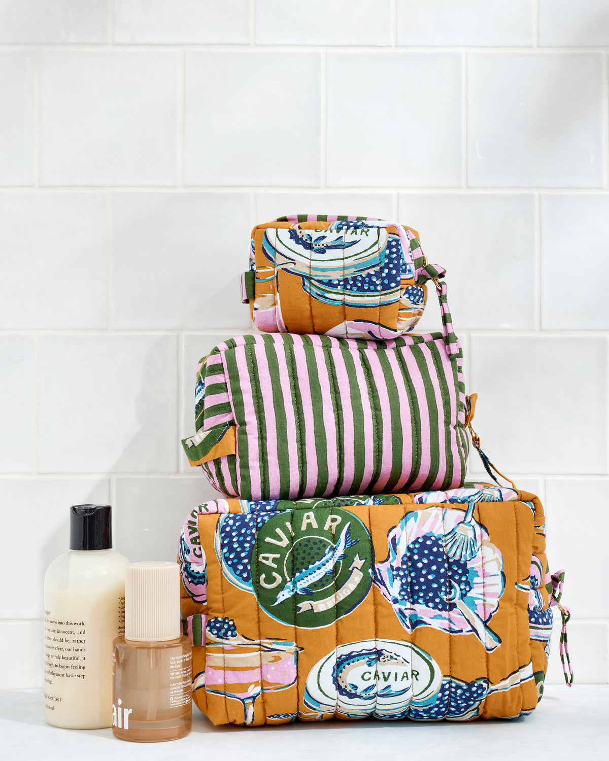 Champagne & Caviar three-pouch set in Goldenrod displayed stacked beside two toiletry bottles.