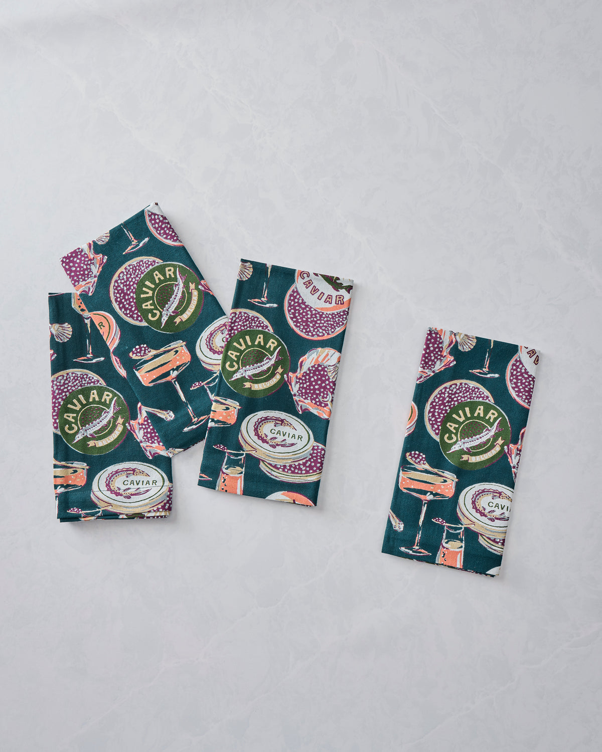 Three folded Champagne and Caviar dinner napkins arranged on a light surface, showing the dark teal caviar and champagne pattern.