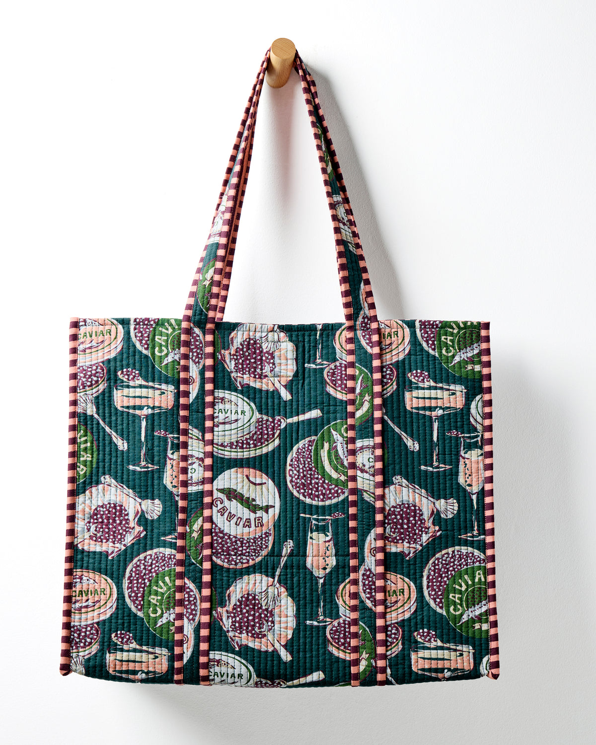 Peacock-feather traveler tote with long shoulder straps and a Champagne & Caviar print, hanging on a wall.
