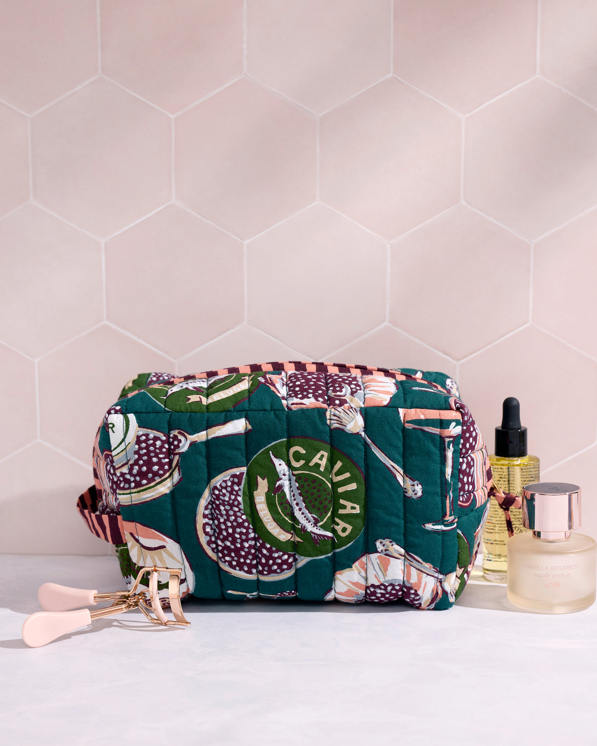Quilted peacock-feather pouch with a Champagne & Caviar print, shown beside cosmetics and an eyelash curler.