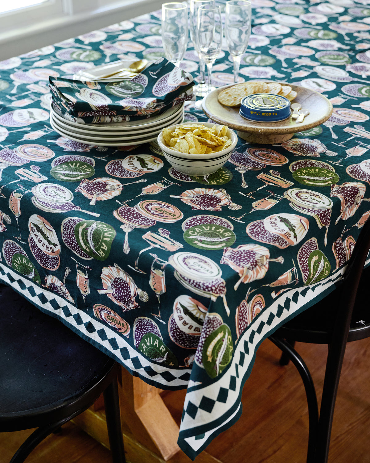 Champagne & Caviar square tablecloth covering a dining table set with plates, glasses, napkins, crackers, and a caviar tin.