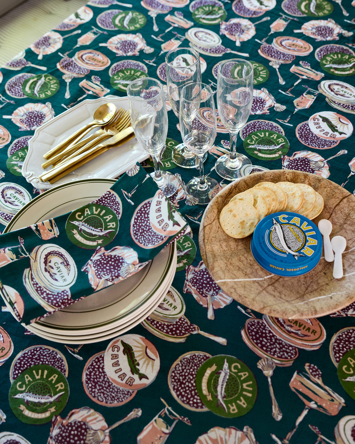 Close view of the Champagne & Caviar square tablecloth with printed caviar tins and seafood motifs beneath plates, utensils, and champagne flutes.