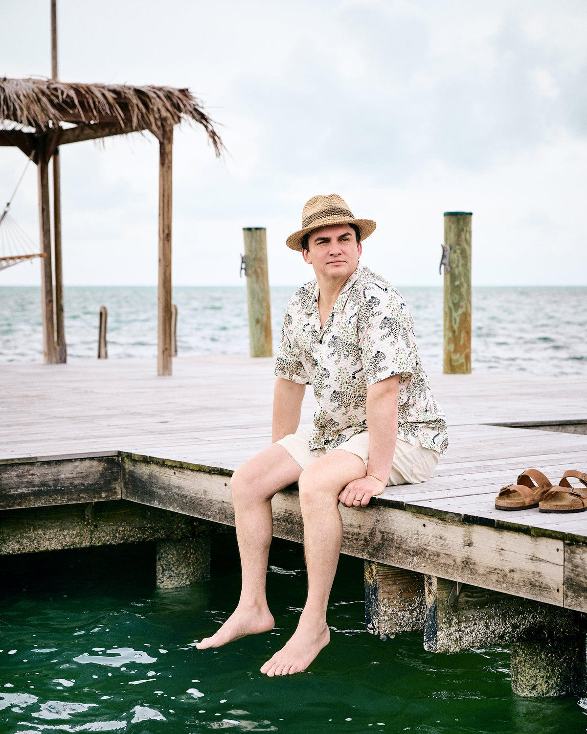 Model seated on a dock wearing the Wedding Cake Bagheera camp shirt, straw hat, and shorts