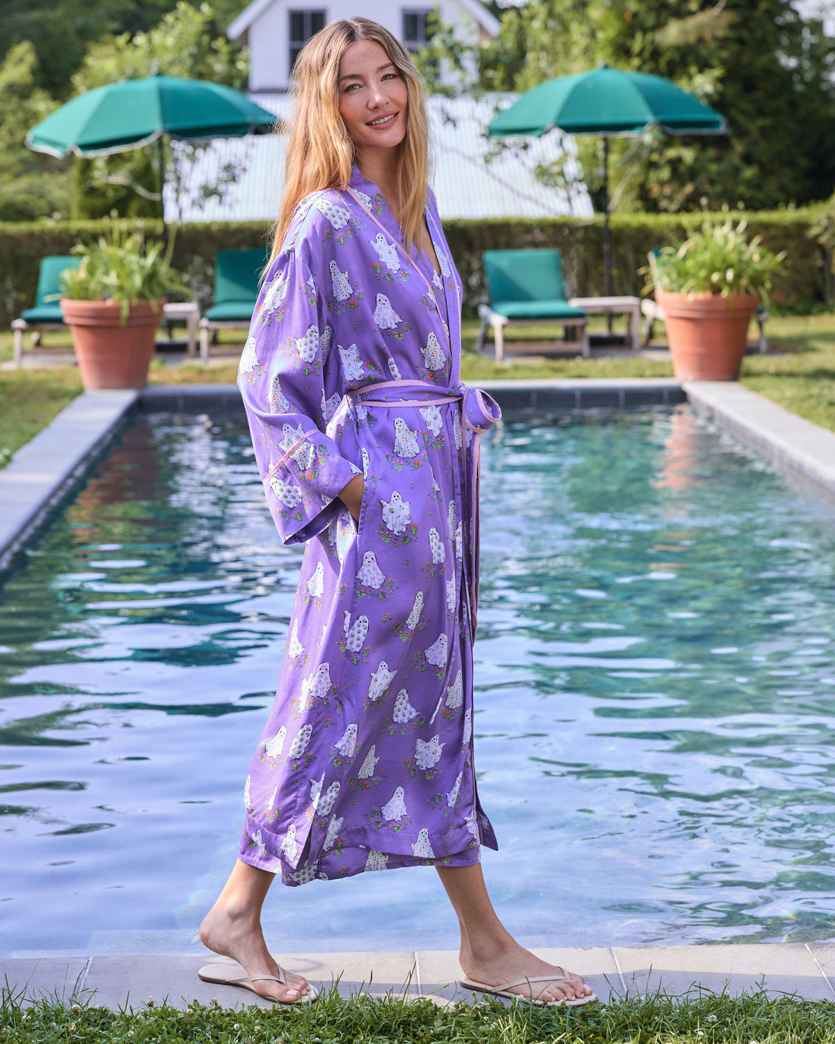 Model wears a long purple robe with a ghost-and-floral print, matching belt, and pockets beside a swimming pool.