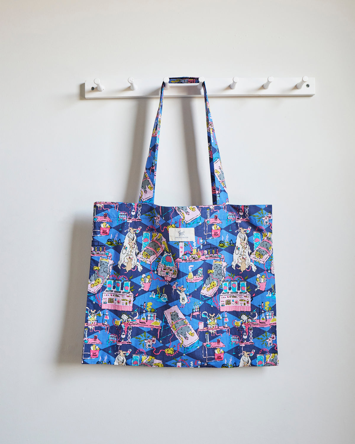 Blue printed tote bag hanging from a white wall rack, showing colorful cat-and-mouse laboratory scenes.
