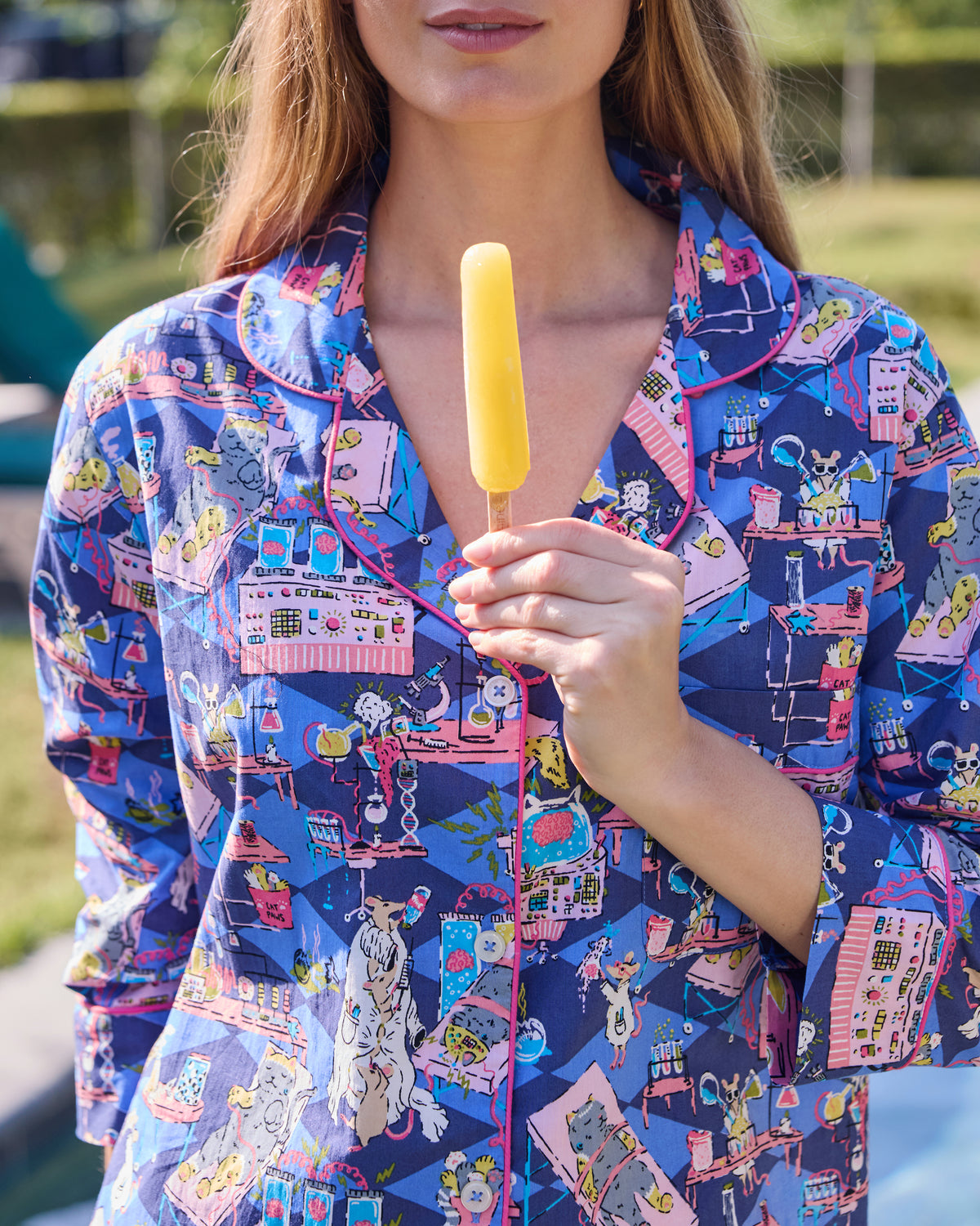 Close view of a blue button-down sleep shirt with a colorful mice-and-laboratory print, while a person holds a yellow popsicle.