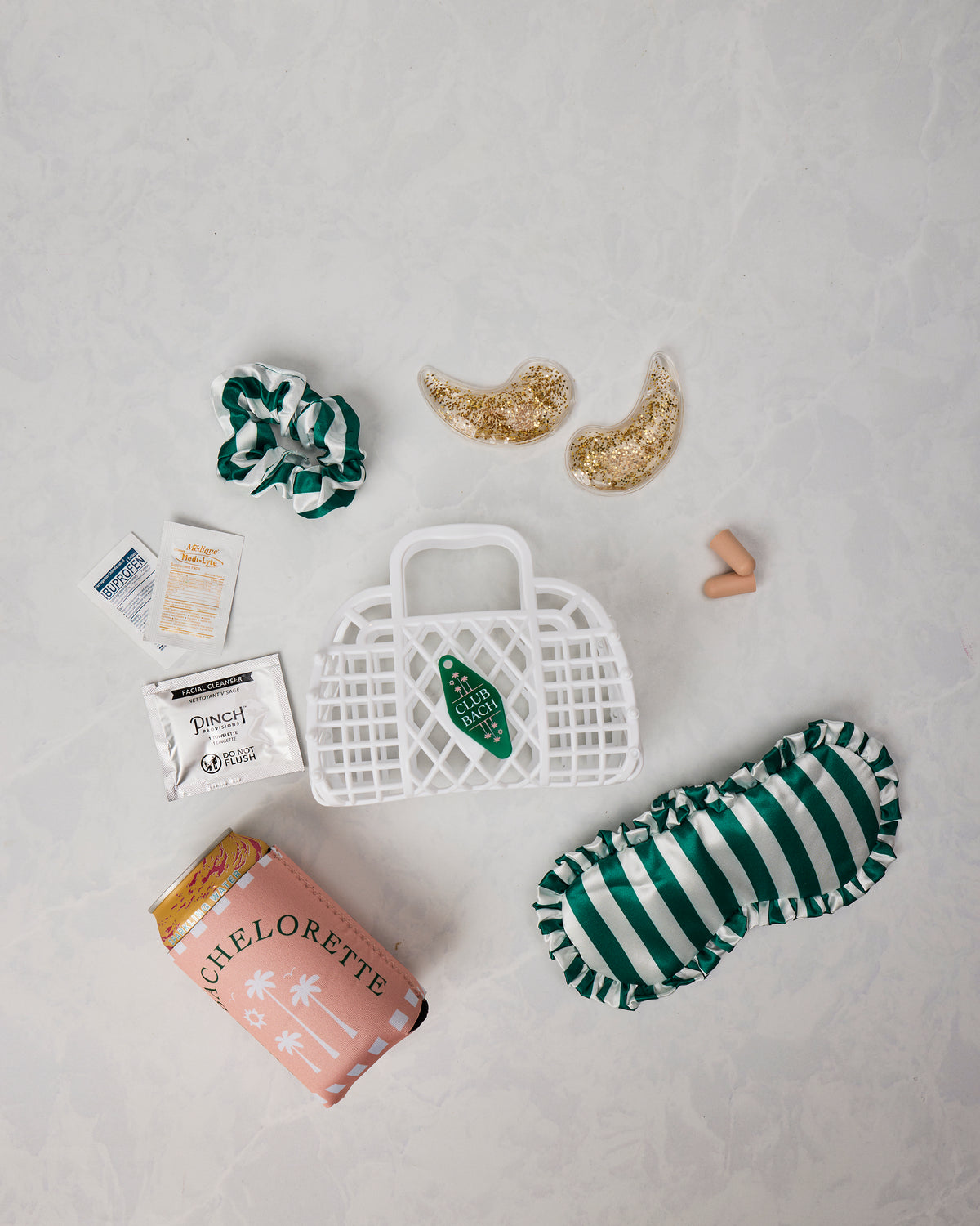 Bachelorette kit contents arranged around a white basket, including a striped sleep mask, striped scrunchies, under-eye patches, earplugs, packets, and pink packaging.