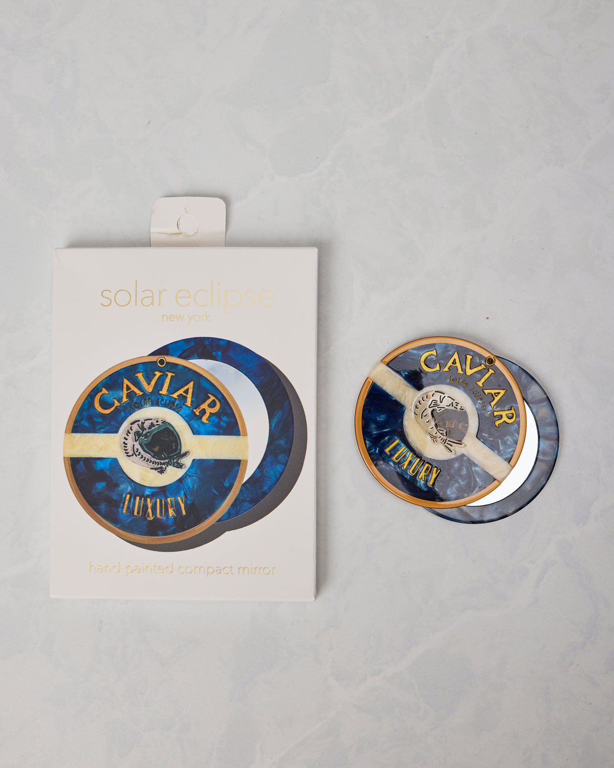 Blue round compact mirror beside its Solar Eclipse New York package, labeled “CAVIAR LUXURY.”