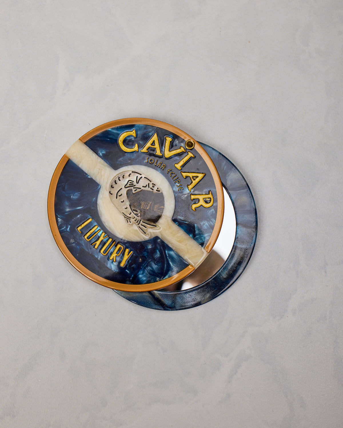 Open blue round compact mirror showing the reflective interior and decorated outer lid labeled “CAVIAR LUXURY.”