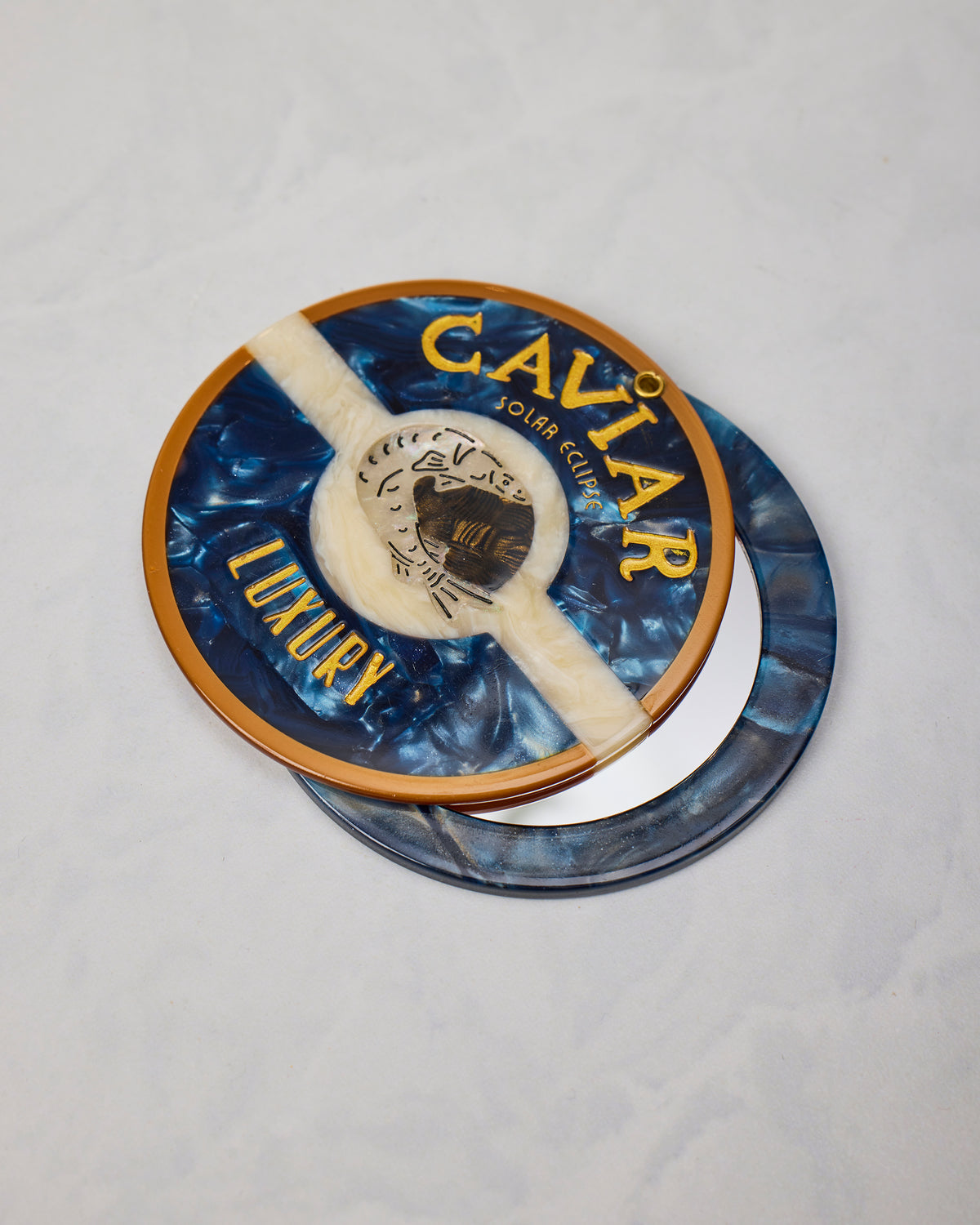 Open blue round compact mirror with the decorated lid resting over the reflective base, labeled “CAVIAR LUXURY.”