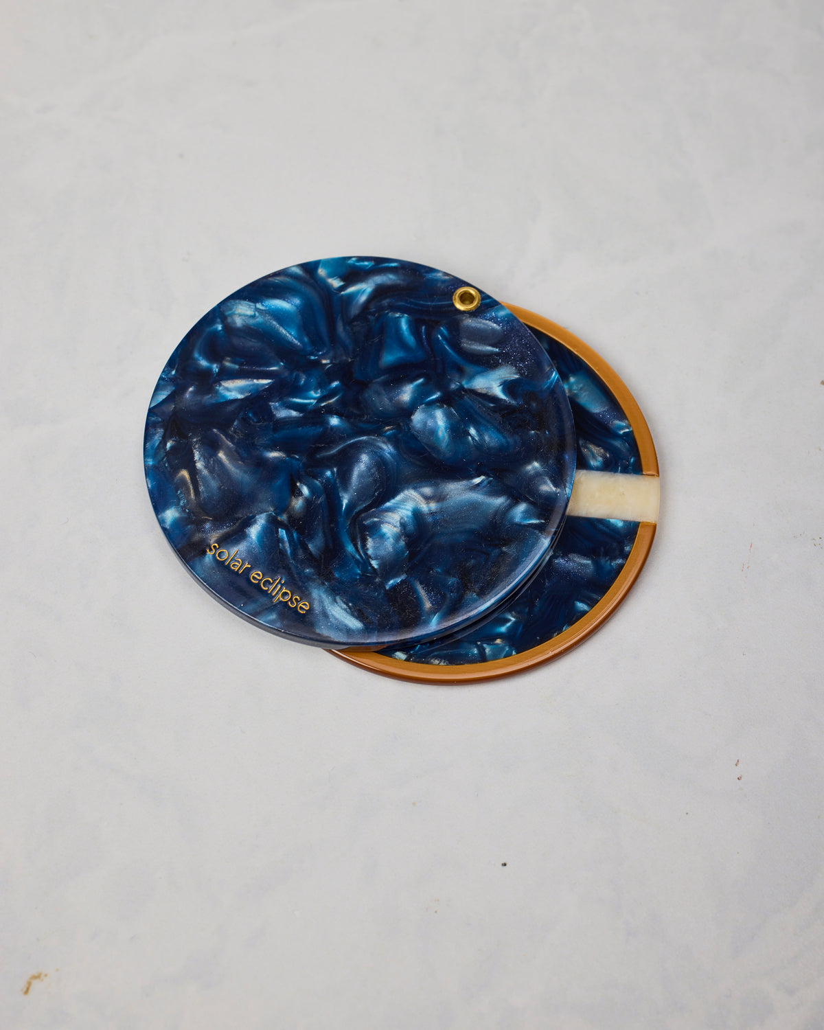 Back of the blue compact mirror showing a marbled blue surface, gold hanging loop, and “Solar eclipse” marking.