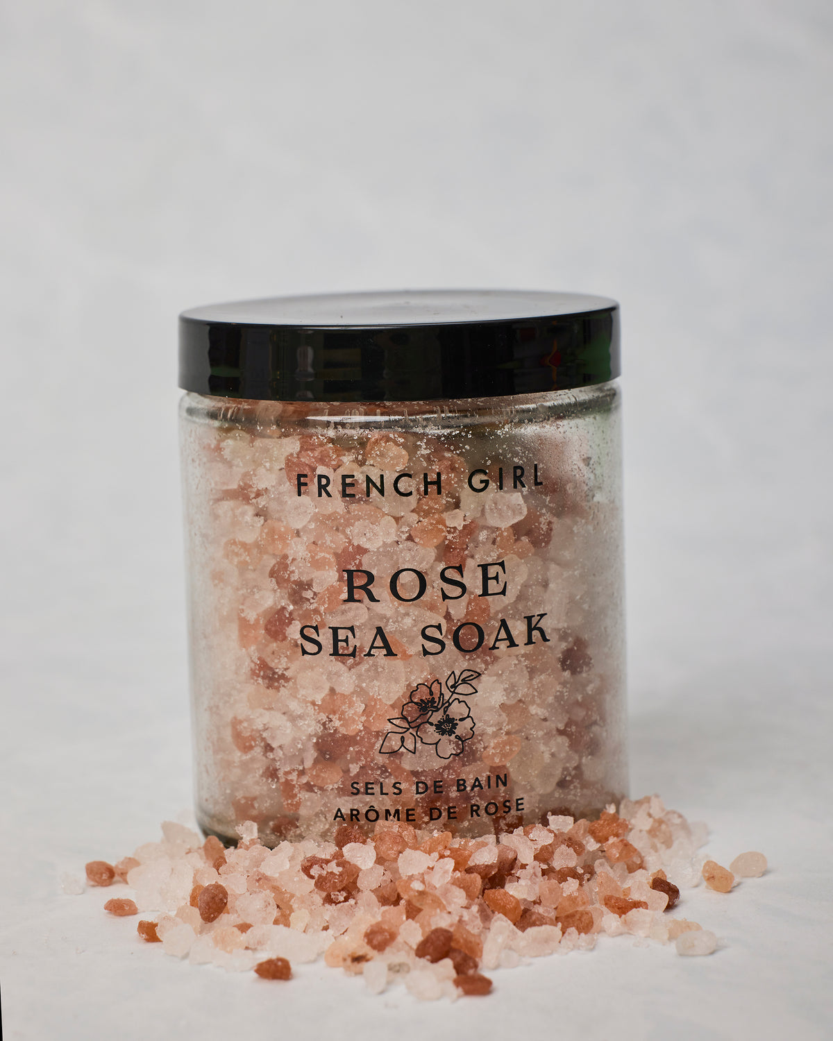 French Girl Rose Sea Soak jar surrounded by loose pink and white bath salts on a light surface.