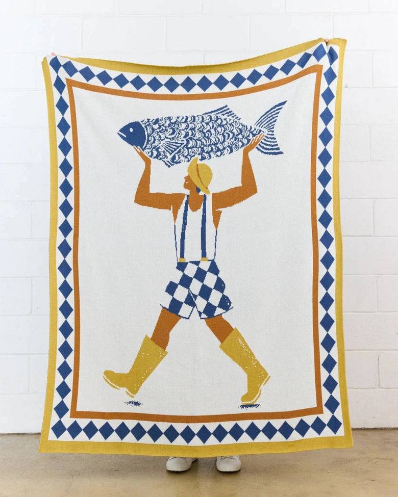 Un Beau Poisson knit throw hanging against a white brick wall, with a figure carrying a large fish.
