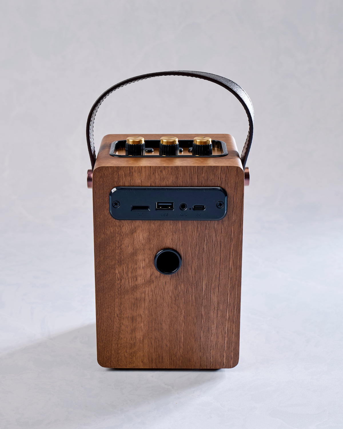 Back view of a brown portable Bluetooth speaker with a curved handle, control knobs on top, connection ports, and a circular rear opening.