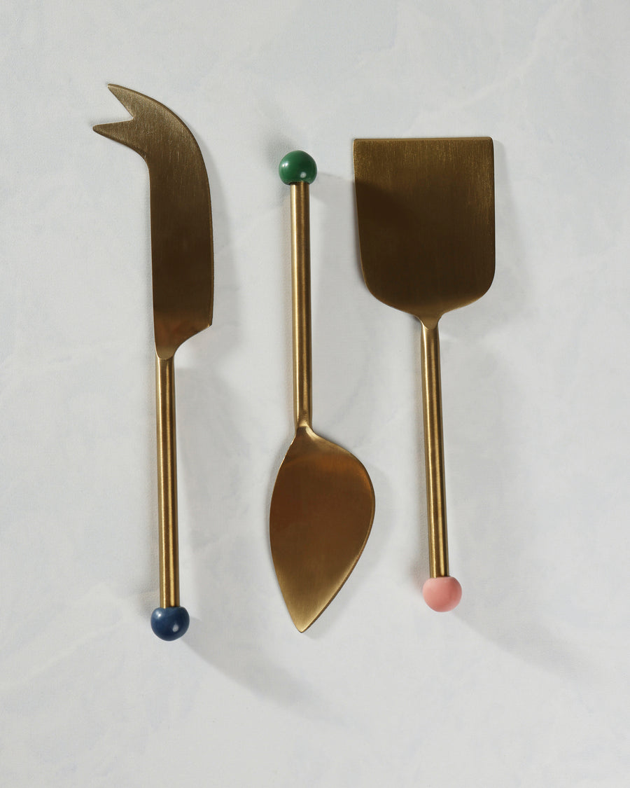 Three-piece gold cheese knife set arranged vertically, including a pointed knife, a spoon-shaped utensil, and a wide flat server.