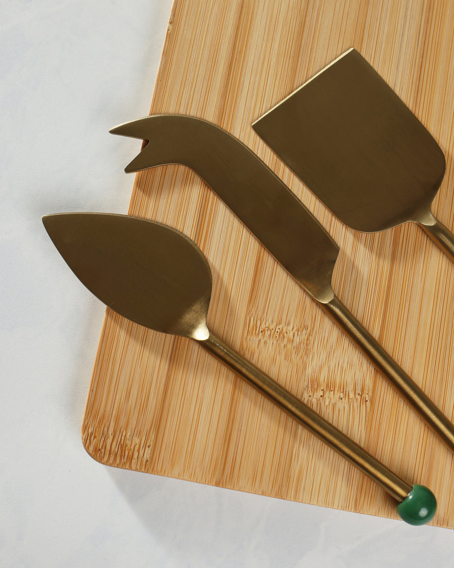 Close angled view of three gold cheese-serving utensils arranged on a bamboo cutting board.