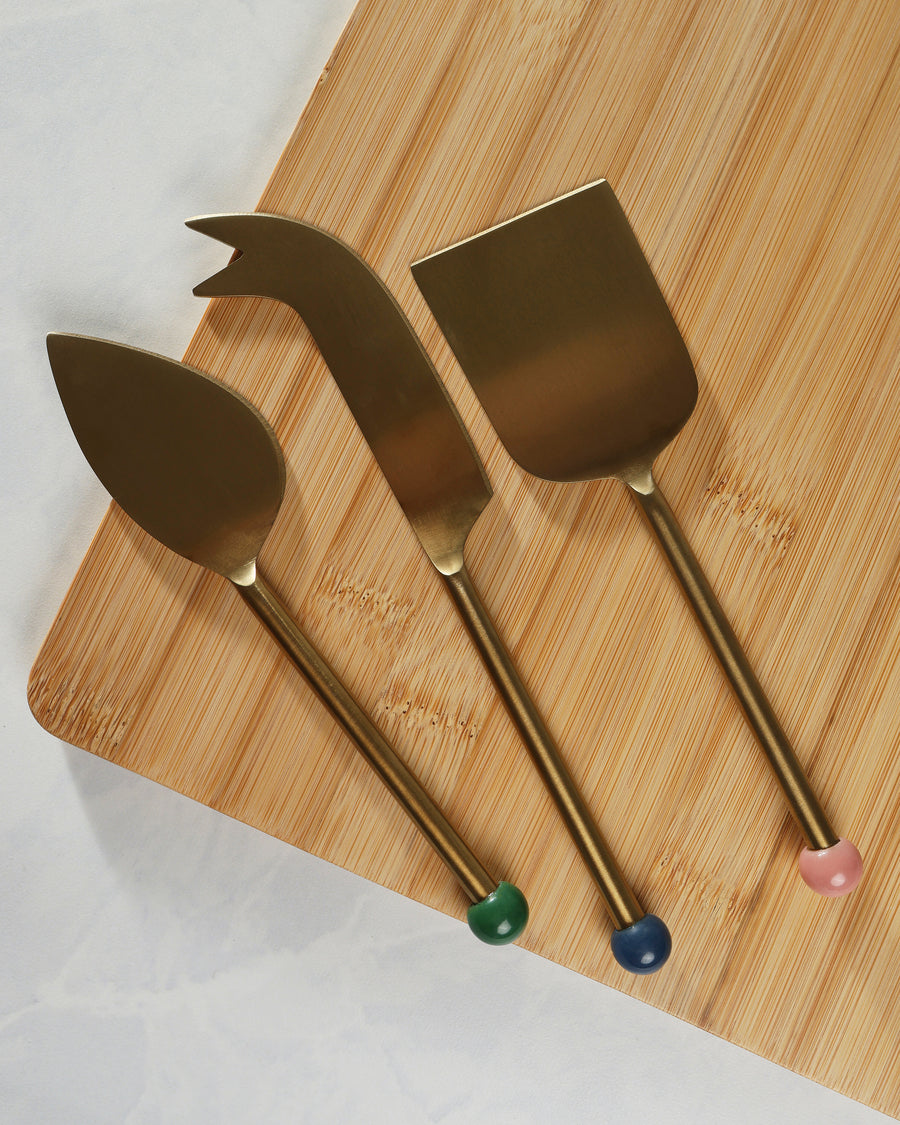 Three gold-finish cheese knives arranged on a bamboo cutting board, including a spreader, server, and fork-tipped knife.
