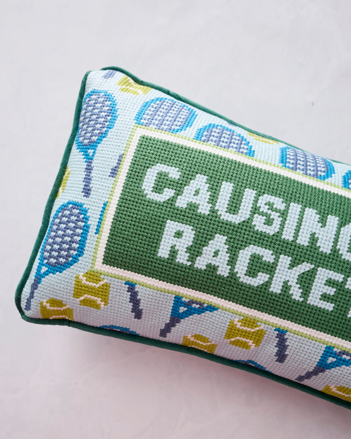 Close-up of a green embroidered needlepoint pillow with a tennis-racket border and the words “CAUSING A RACKET.”