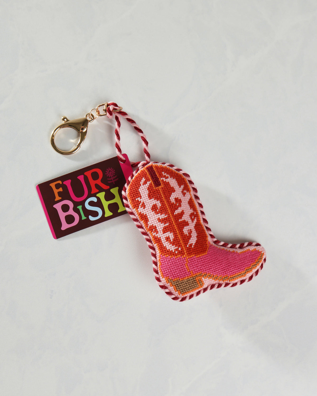 Pink embroidered cowboy boot bag charm with a gold clip and twisted hanging loop, shown beside a FURBISH tag.