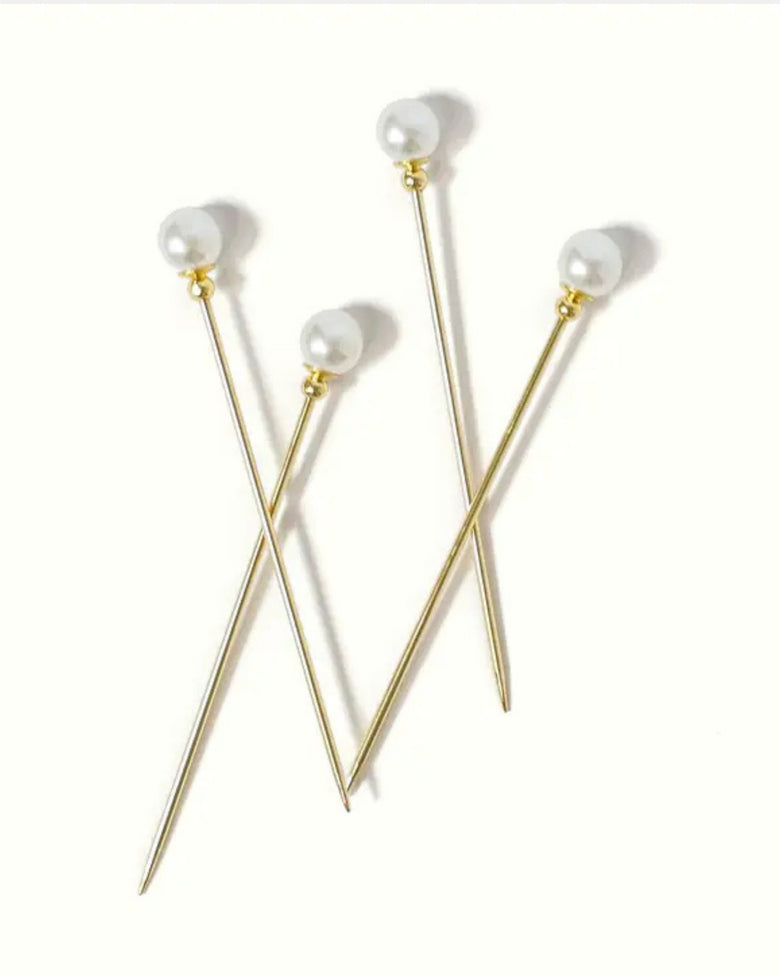 Four gold cocktail picks with white faux-pearl tops arranged on a white background.