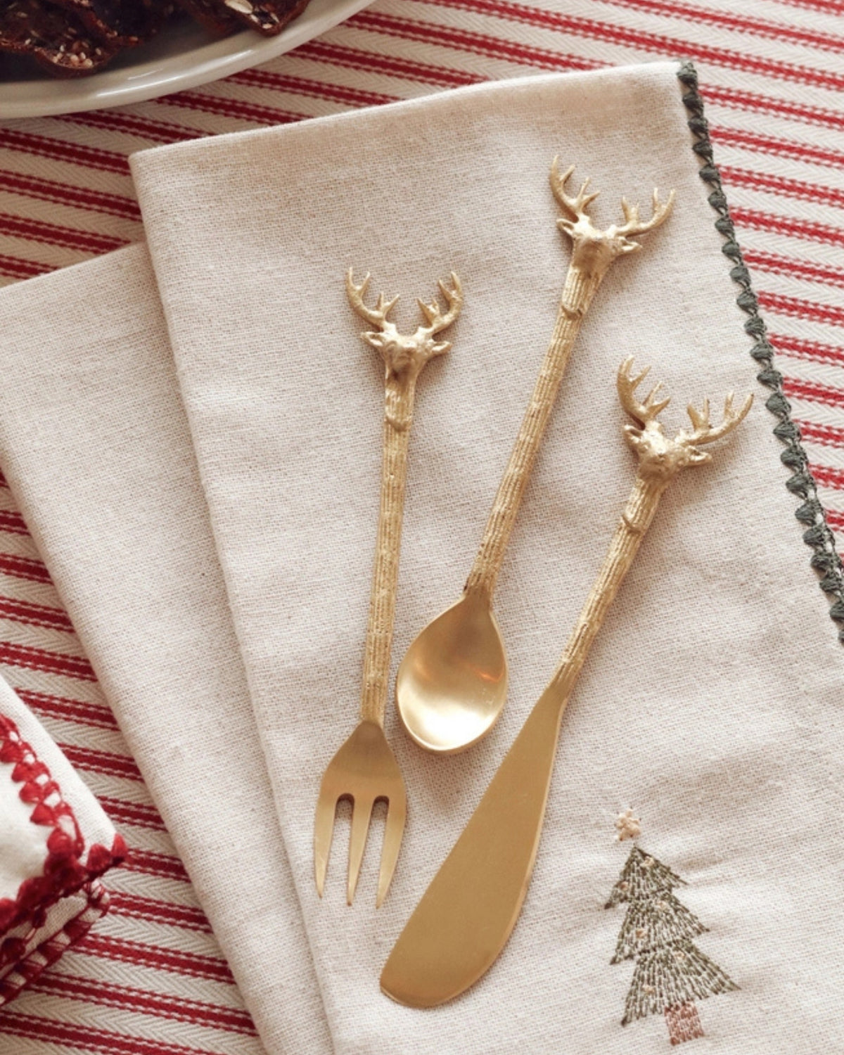 Three gold-colored stag-handled cheese tools arranged on a folded napkin beside a holiday table setting.