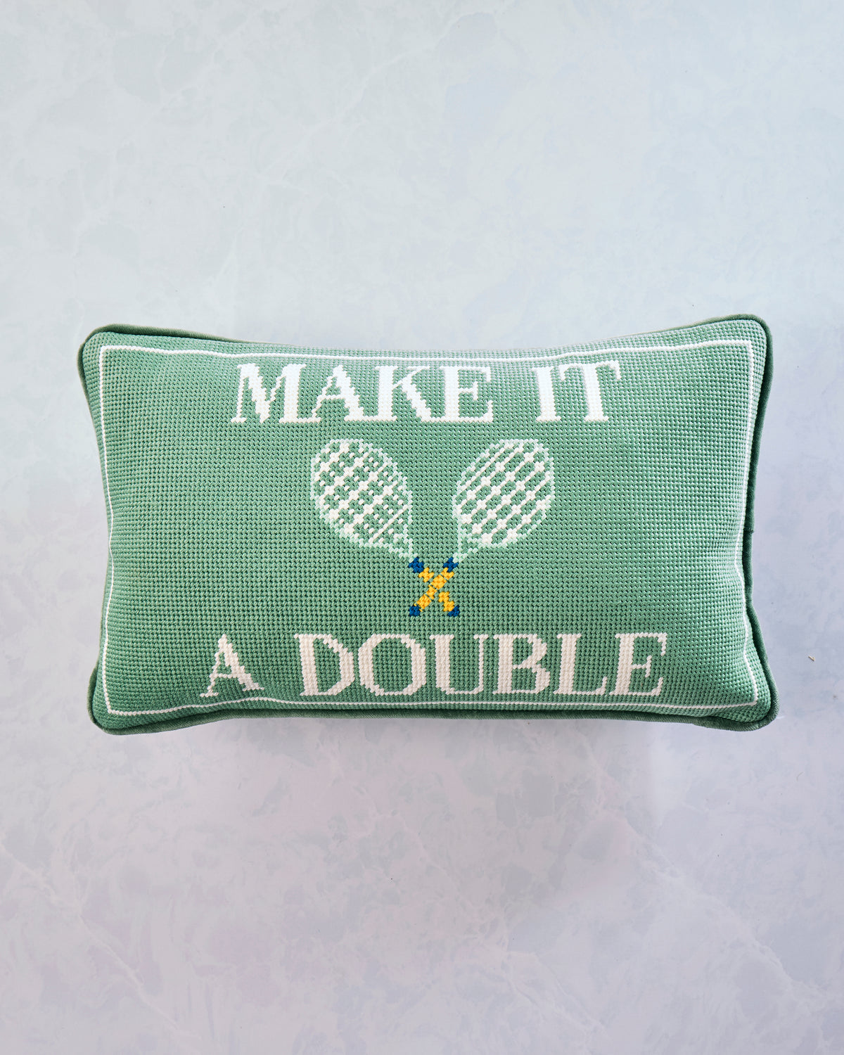 Front of a green needlepoint pillow reading “MAKE IT A DOUBLE” beneath two crossed tennis rackets.