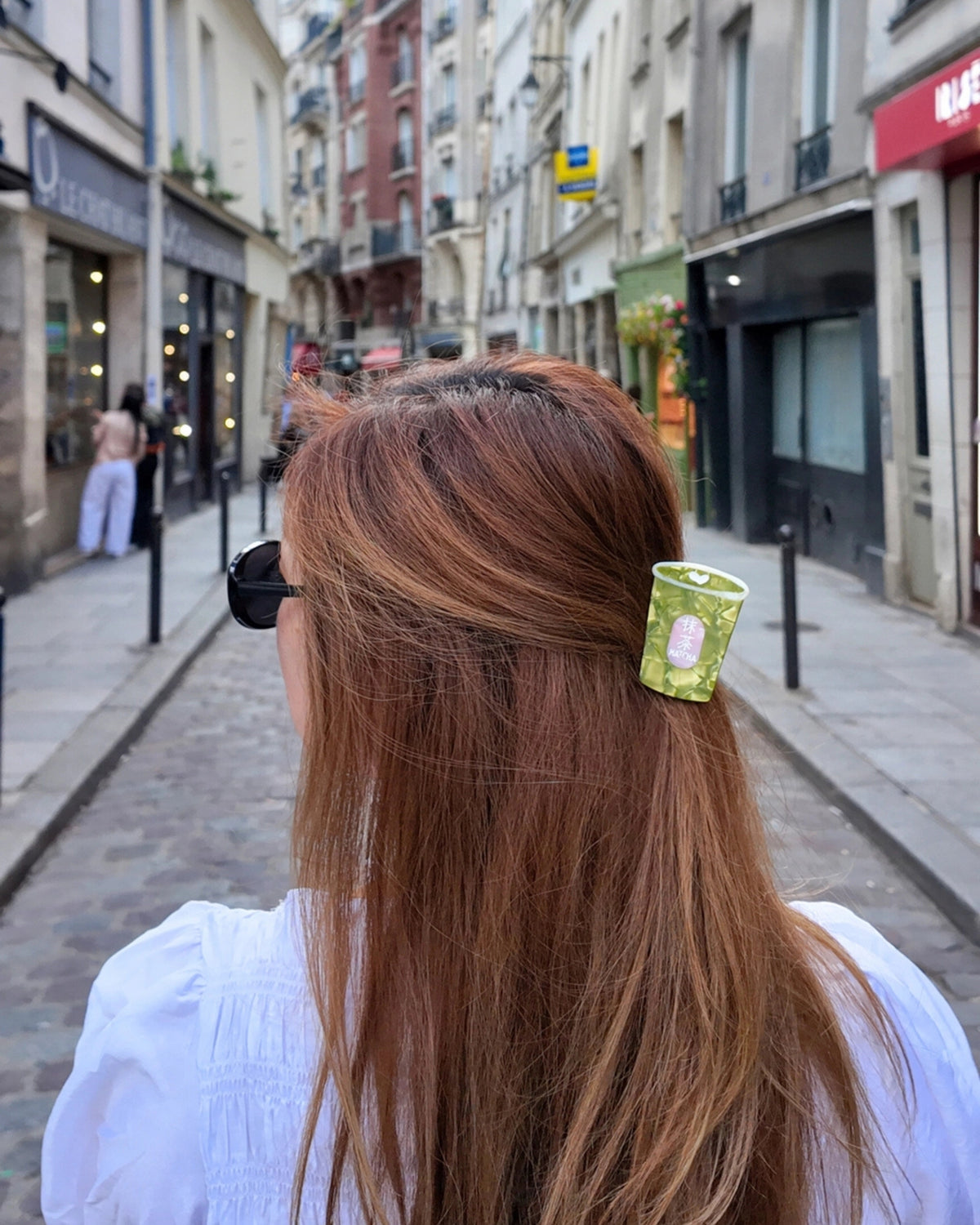 Green mini claw hair clip securing a section of hair, viewed from behind on a city street.