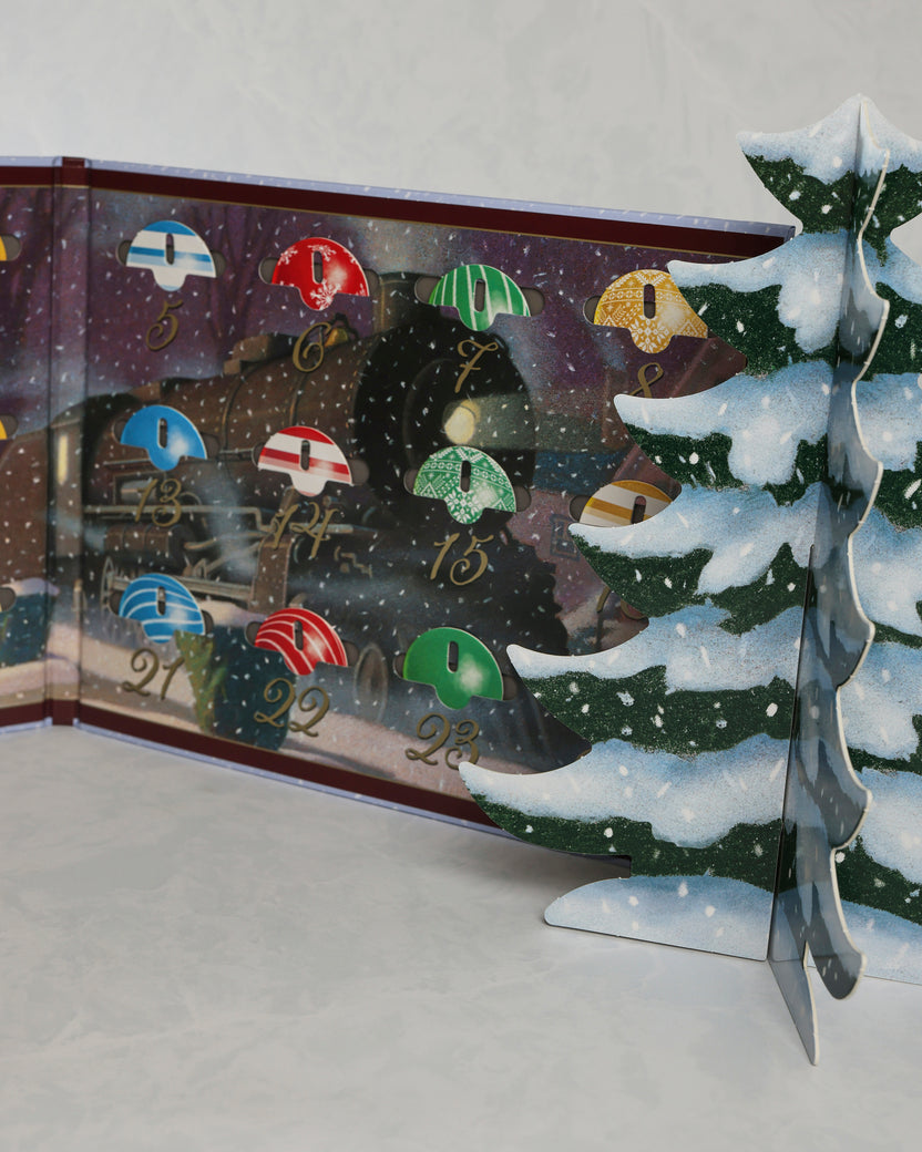 Open Christmas countdown calendar showing numbered ornaments and a freestanding snowy Christmas tree