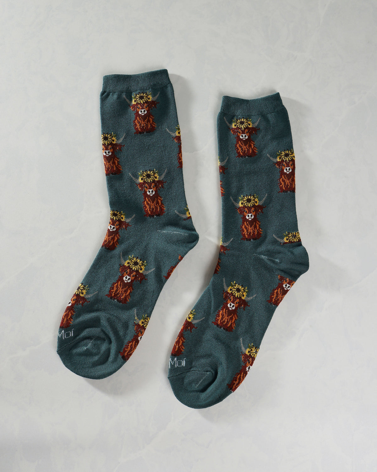 Pair of teal crew socks covered with repeating Highland cow illustrations wearing flower crowns.