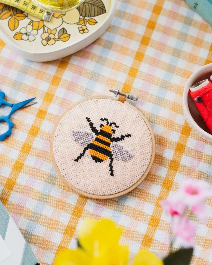 Bumble bee cross-stitch design in a wooden hoop on peach fabric, with craft supplies arranged on a gingham table.