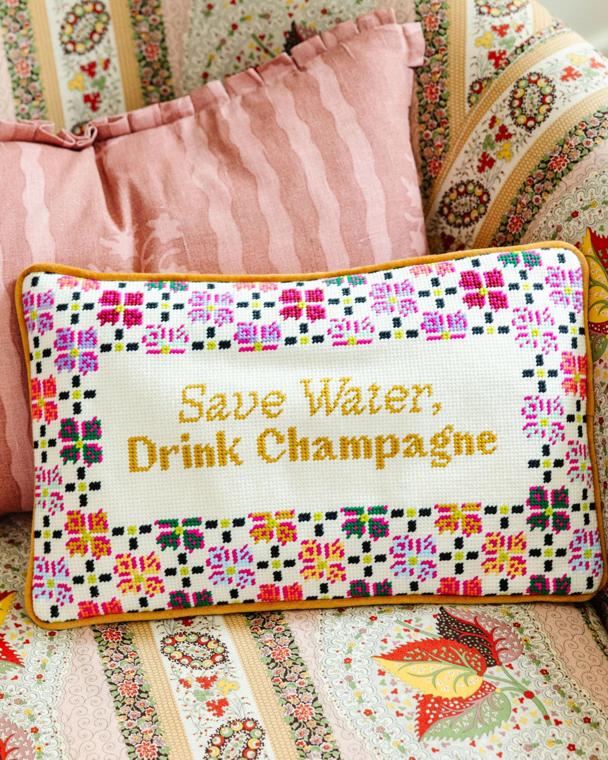 Needlepoint pillow embroidered with “Save Water, Drink Champagne” and a colorful floral border, shown against patterned cushions.