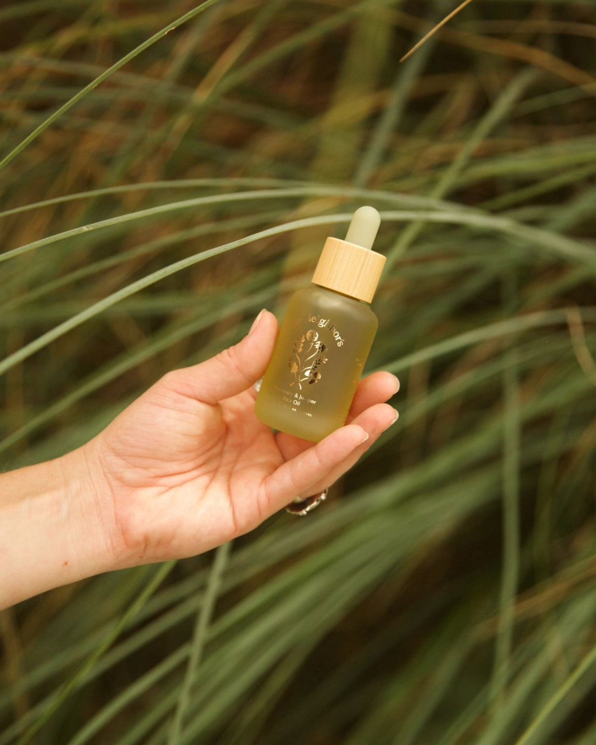 A hand holds the Rosemary & Juniper Hair Oil dropper bottle against a background of green grass.