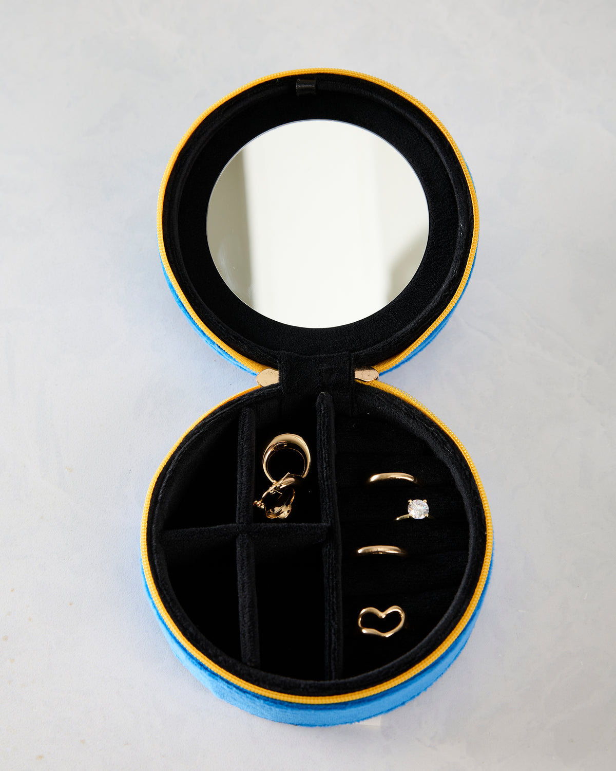 Top view of an open blue velvet jewelry zip case showing its round mirror, divided compartments, and gold-tone jewelry.