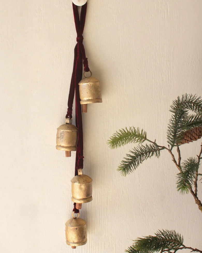Gold jingle bells hanging vertically from a maroon ribbon on a wall beside evergreen branches.