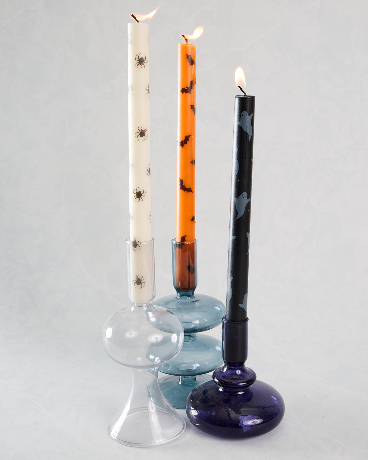 Three lit Halloween taper candles in glass holders: ivory with spiders, orange with bats, and black with ghosts.