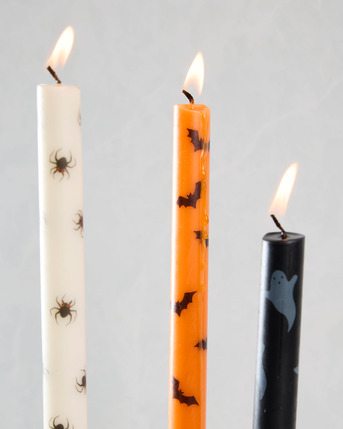 Close-up of lit ivory, orange, and black Halloween taper candles with spider, bat, and ghost designs.