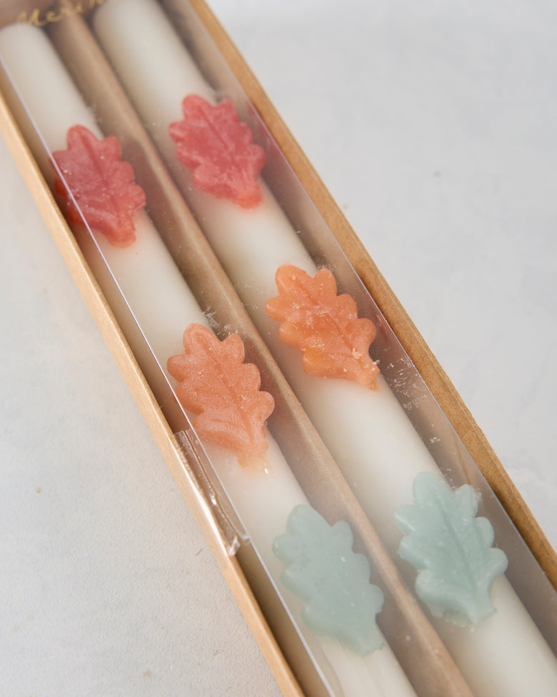 Angled close-up of two white candles in a box, decorated with pink, orange, and pale green leaf-shaped accents.
