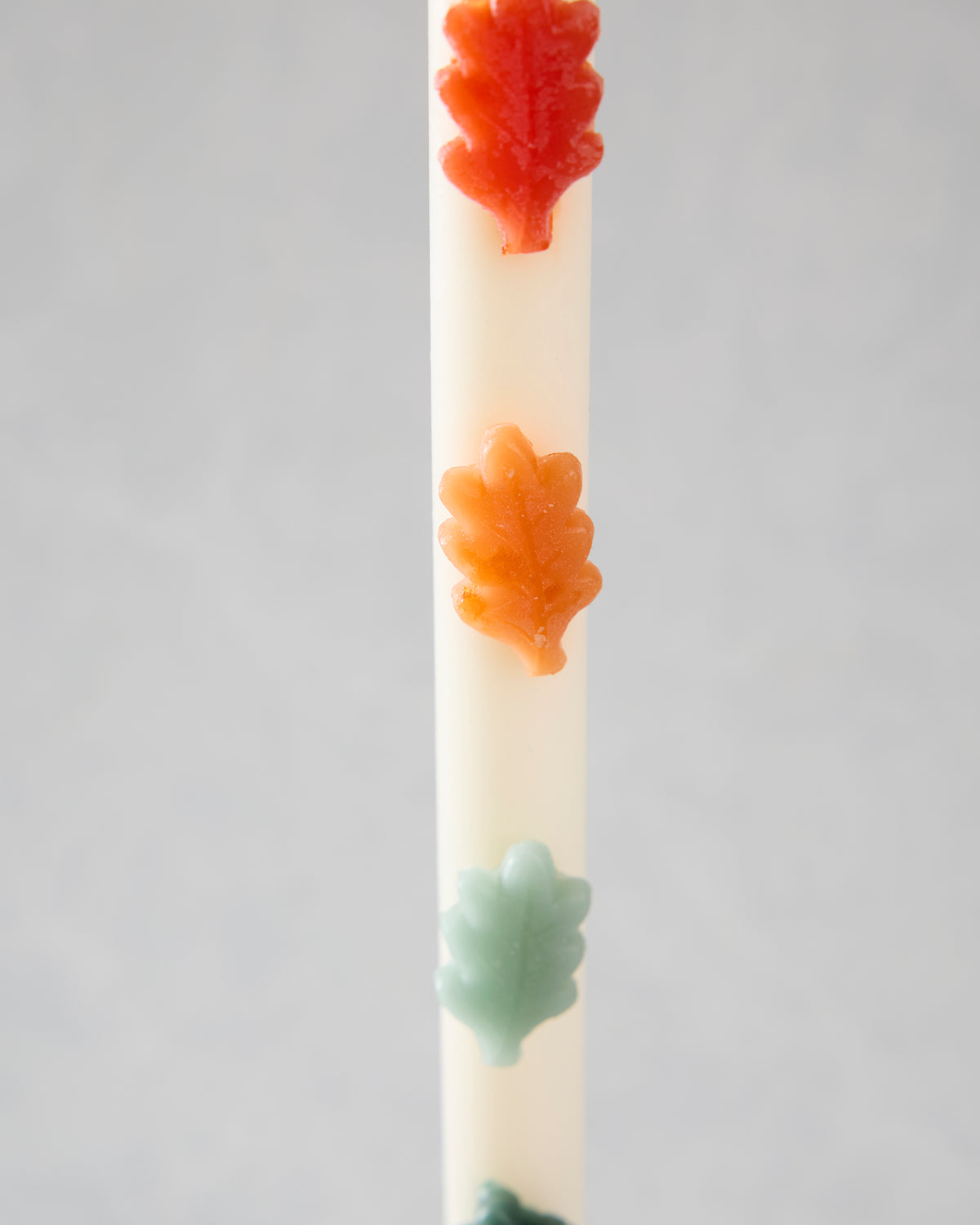 Close-up of a white candle decorated with red-orange, orange, pale green, and dark green leaf-shaped accents.