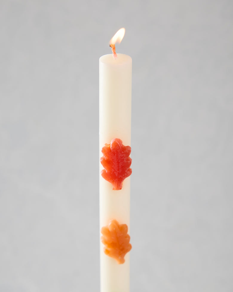 Lit white leaf-decorated candle with red-orange and orange leaf-shaped accents.