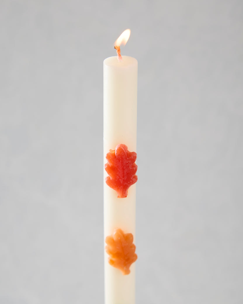 Lit white leaf-decorated candle with red-orange and orange leaf-shaped accents.