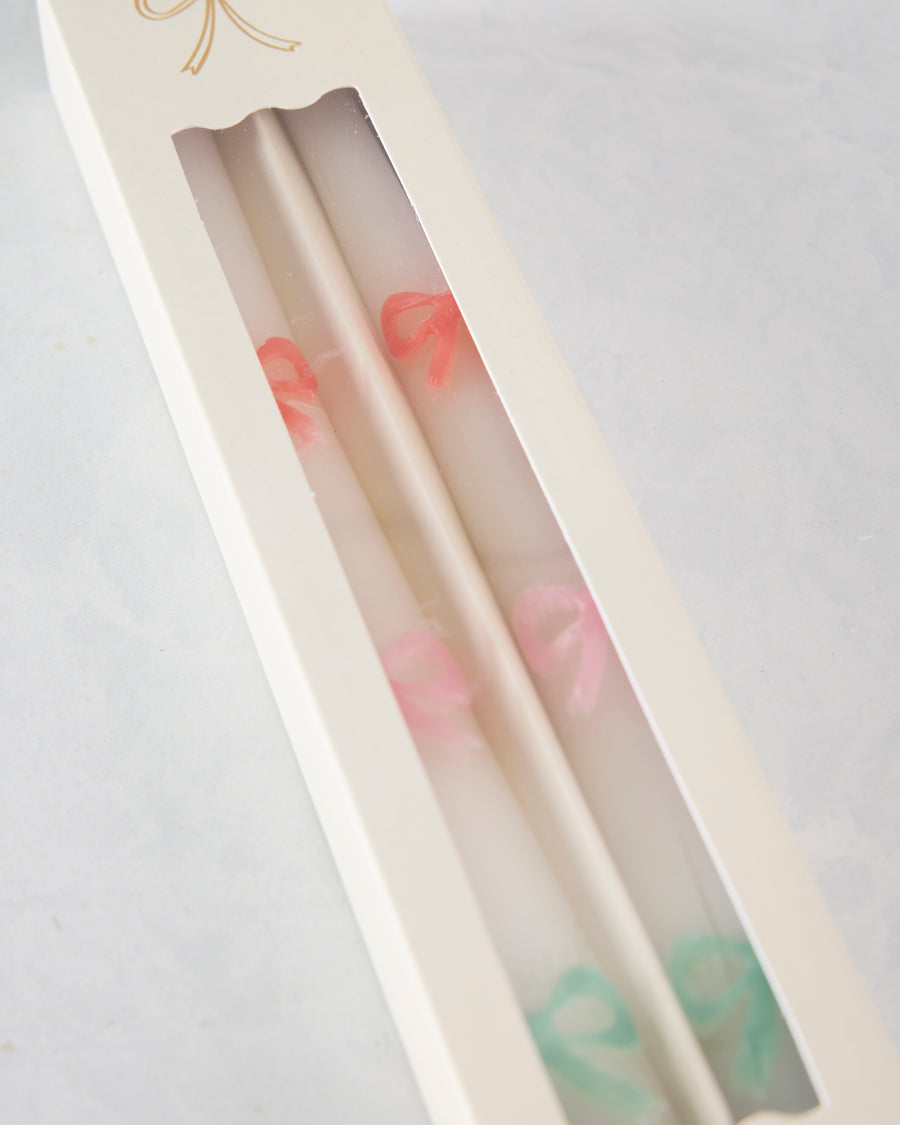 Angled close-up of two white taper candles in a windowed box, showing orange, pink, and green bow details.