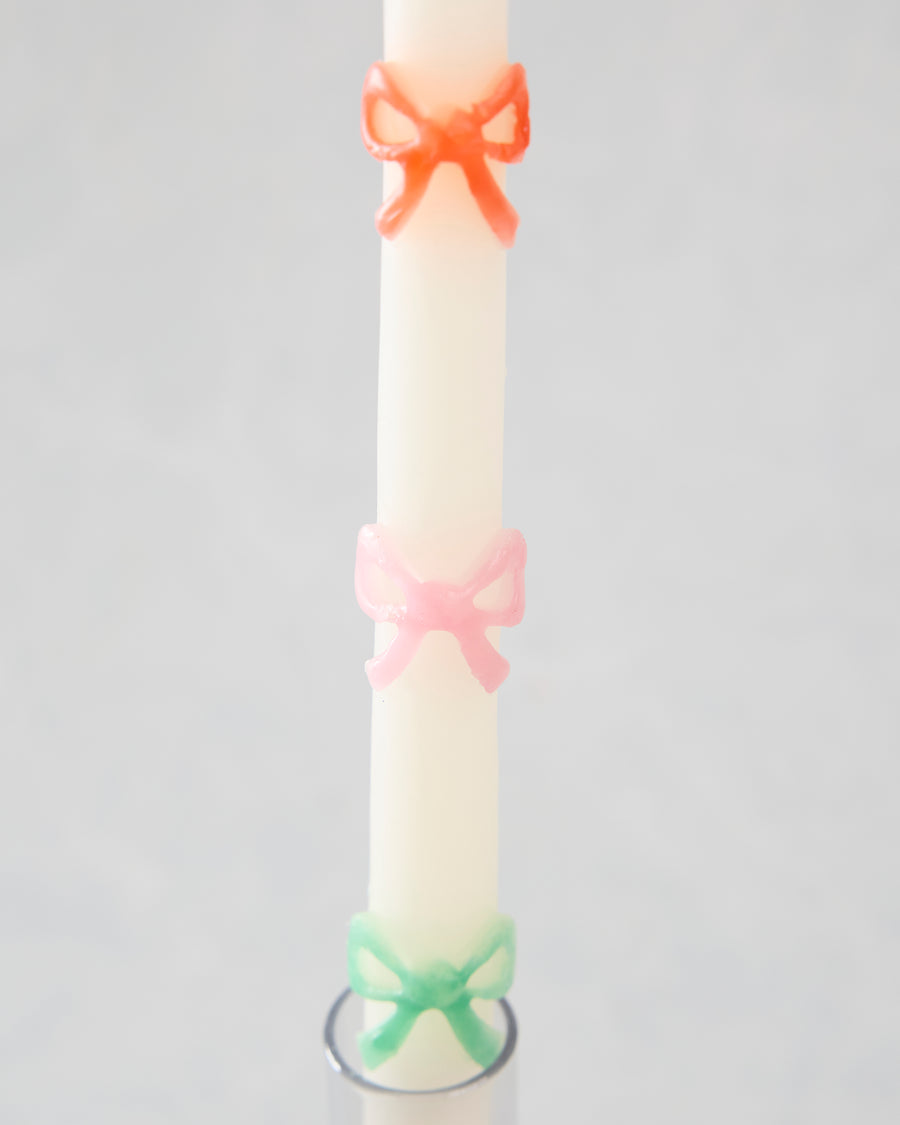 Close-up of a white taper candle with orange, pink, and green bow decorations above a candle holder.