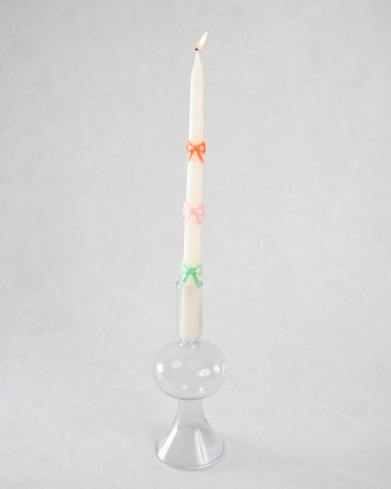Lit white taper candle with orange, pink, and green bow decorations displayed in a clear glass holder.