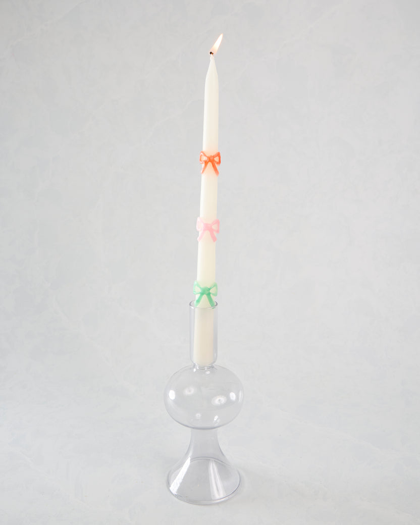 Lit white taper candle with orange, pink, and green bow decorations displayed in a clear glass holder.