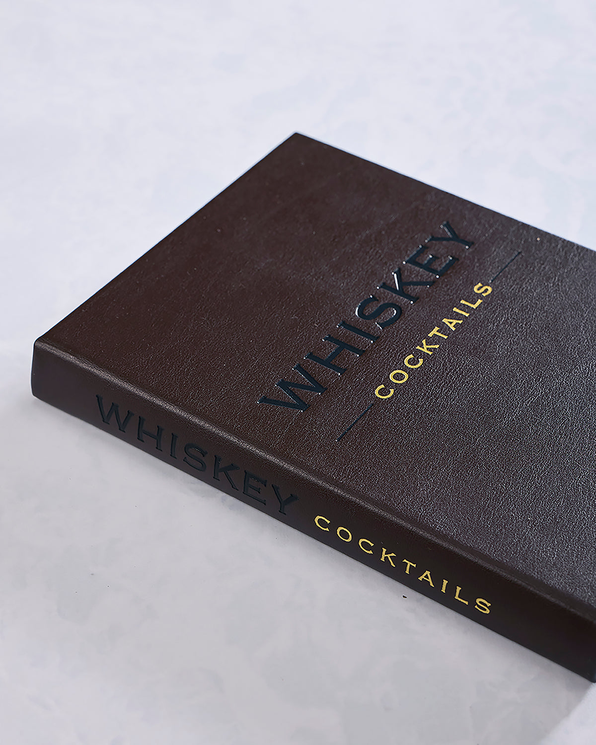 Closed dark brown Whiskey Cocktails book photographed at an angle, with black and gold lettering on the cover and spine.