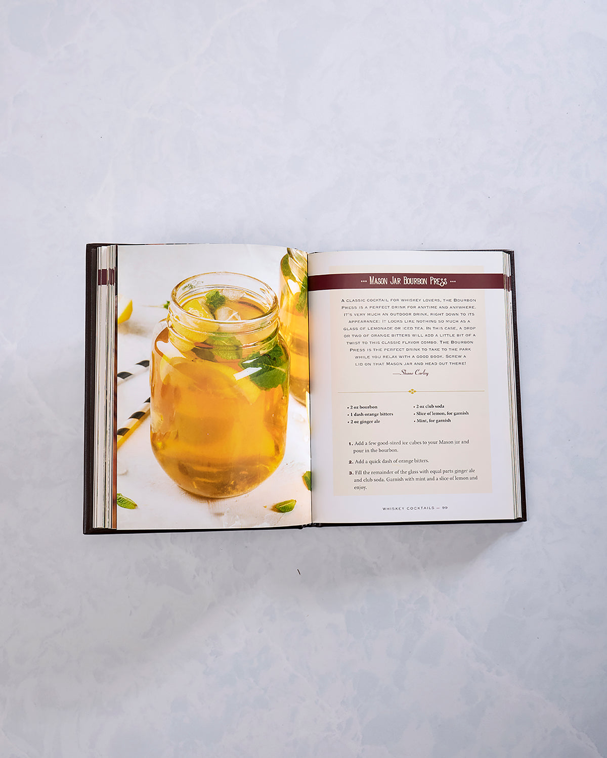 Open whiskey cocktail book showing a drink in a mason jar beside a recipe page titled “Mason Jar Bourbon Press.”