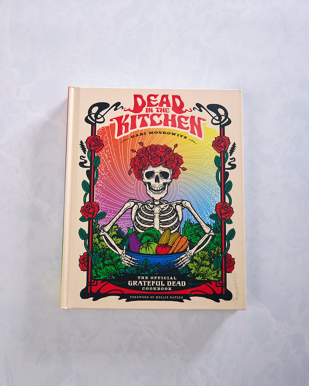 Front cover of the cookbook “Dead in the Kitchen,” featuring a skeleton with red roses holding a bowl of vegetables.